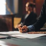 Person writing at desk with documents and glass.