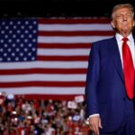 Donald Trump in suit stands before large American flag