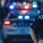OUTRAGE: Nails KILL Hero Police Dog Police officer stands near patrol car with lights flashing.