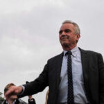 RFK Jr. to ILLEGALS – NO MORE! Robert F Kennedy Jr in a suit stands outdoors cloudy sky background