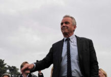 Robert F Kennedy Jr in a suit stands outdoors cloudy sky background
