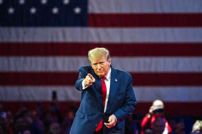 shutterstock_2455396913.jpg Donald Trump pointing with large American flag backdrop.