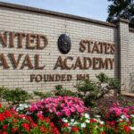 Purge ACCELERATES — Academy Commander OUT Sign of the United States Naval Academy with flowers in front