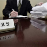 IRS Chaos Looms: Massive Staff Cuts IRS sign on desk, person handling paperwork behind.