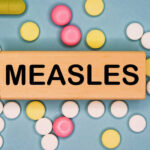 Explosive Measles Spread: Famed College on Edge Wooden block with the word 'MEASLES' surrounded by various pills