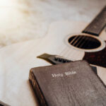 An acoustic guitar next to a Holy Bible on a soft surface