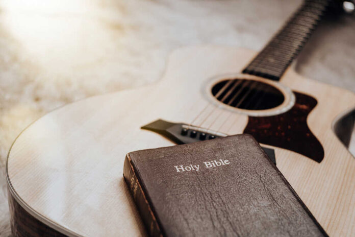 An acoustic guitar next to a Holy Bible on a soft surface