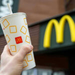 McDonald’s Enjoying Comeback Nobody Expected Hand holding McDonalds cup in front of restaurant sign.