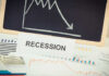 A sign reading 'RECESSION' in front of various financial charts and a downward trend graph