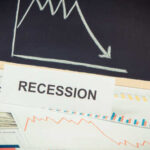 A sign reading 'RECESSION' in front of various financial charts and a downward trend graph