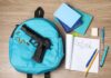 A blue backpack with a handgun and school supplies on a wooden surface