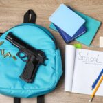 A blue backpack with a handgun and school supplies on a wooden surface