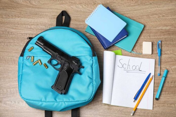 shutterstock_2483467309.jpg A blue backpack with a handgun and school supplies on a wooden surface
