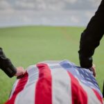 Two individuals standing beside a flag-draped casket in a green field