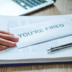 A hand resting on an envelope labeled 'YOU'RE FIRED' on a desk with papers and a pen
