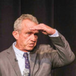 RFK Jr. WARNS U.S. Hospitals Robert F Kennedy in gray suit shielding eyes.