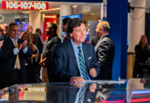 People around news desk, Tucker Carlson in suit at the center.