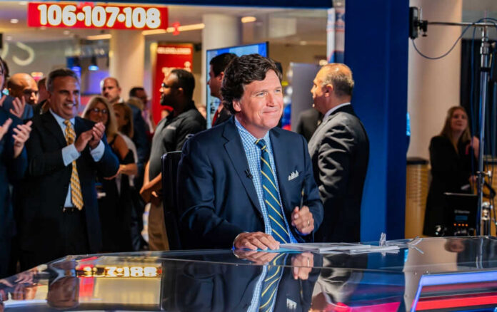 People around news desk, Tucker Carlson in suit at the center.