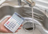Water Bills EXPLODE — Families Crushed Nationwide A hand holding a calculator above a kitchen sink with running water