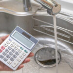 Water Bills EXPLODE — Families Crushed Nationwide A hand holding a calculator above a kitchen sink with running water