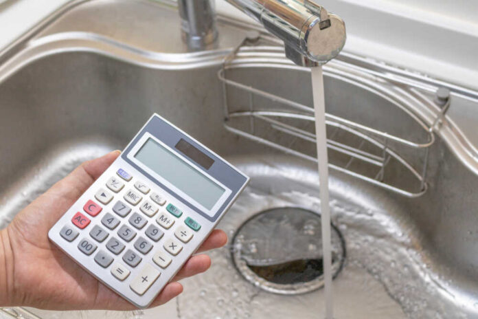 A hand holding a calculator above a kitchen sink with running water