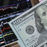 A close-up of a hundred-dollar bill in front of stock market graphs