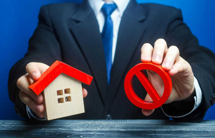 shutterstock_1679810578 Businessman holding house model and prohibition sign