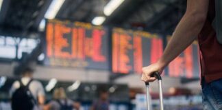 A traveler holding a suitcase in an airport terminal with a flight information display in the background
