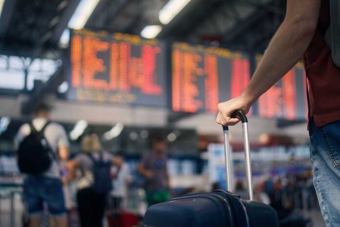 Traveling,By,Airplane.,Man,Waiting,In,Airport,Terminal.,Selective,Focus A traveler holding a suitcase in an airport terminal with a flight information display in the background
