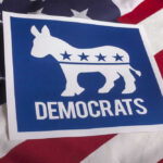 VIDEO: Armed Rush At Dem Candidate Stuns Crowd Democrats logo on American flag background.