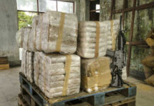 HUGE: Narco-Terror Boss Now in U.S. Stacked packages wrapped in plastic next to a rifle in a warehouse setting