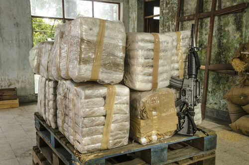 Drug,Warehouse,This,Scene,Was,Staged,To,Recreate,An,Illegal Stacked packages wrapped in plastic next to a rifle in a warehouse setting