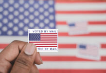 Mystery Voter List Sparks Legal Threat Hand holding a vote-by-mail sticker with U.S. flag