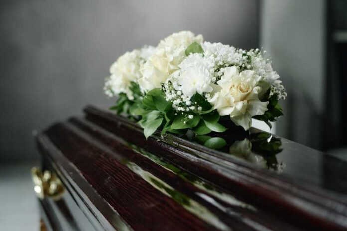 Close,Up,Of,Dark,Wooden,Coffin,Adorned,With,Roses,And A floral arrangement of white roses and greenery on a polished casket